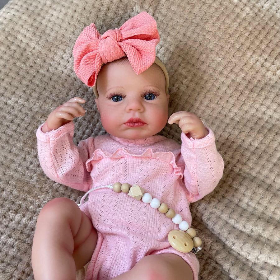 Daphne, a 20 inch awake reborn baby girl with blue eyes, wearing a pink romper and big pink bow