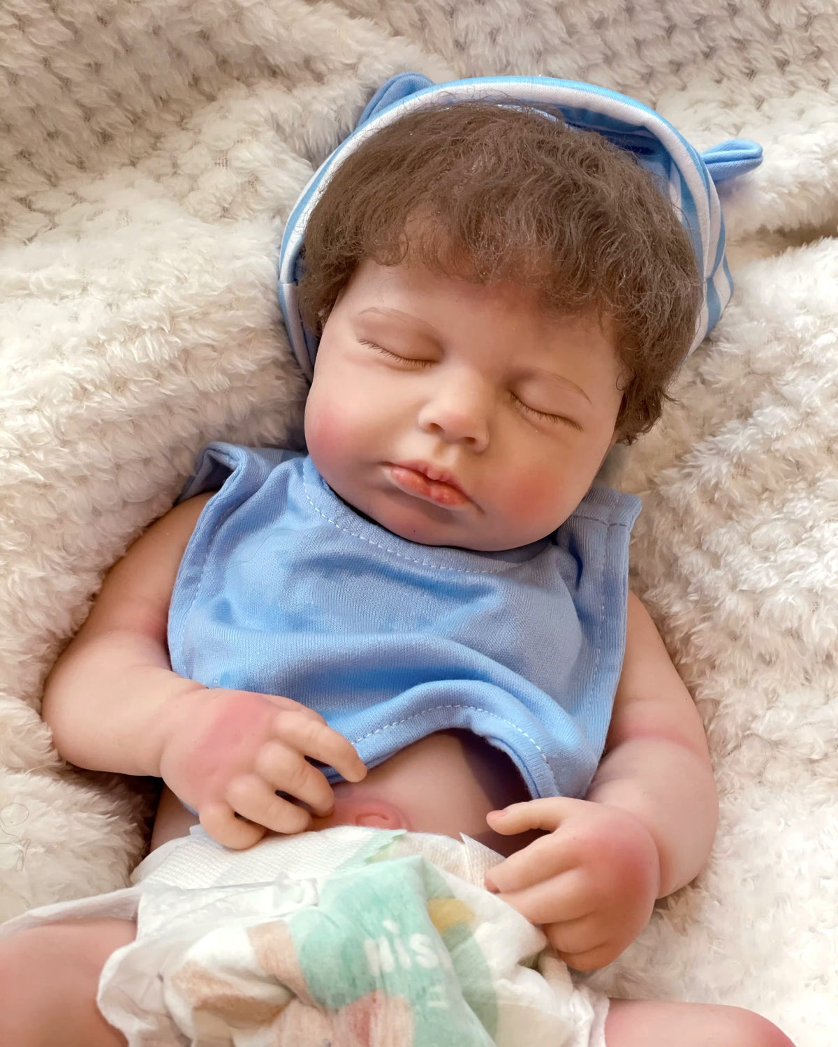 LouLou, a full silicone reborn baby boy with rooted brown hair, sleeping on a cream blanket